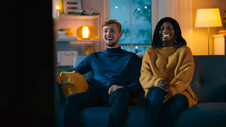 Happy Diverse Young Couple Watching Comedy on TV while Sitting on a Couch, they Laugh and Enjoy...