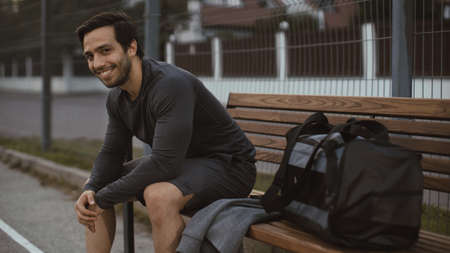 Athletic Young Man in Sports Outfit is Sitiing on a Bench and Looking into Camera. He will Jogging in the Street. Urban Environment.の写真素材