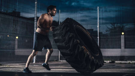 Strong Muscular Fit Young Shirtless Man is Doing Exercises in a Fenced Outdoor Basketball Court. Hes Flipping a Big Heavy Tire in an Afternoon After Rain in a Residential Neighborhood Area.の写真素材