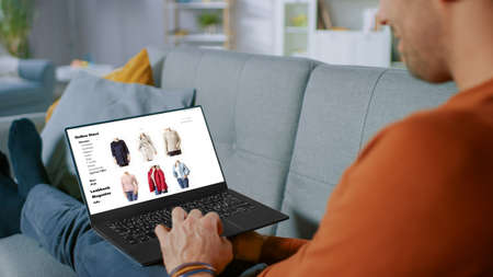 Handsome Young Man Sits at Home Uses Laptop and Browses Through Online Retailer Clothes Selling Store. Man Does Online Shopping for Clothing Items as a Gift for His Wife.の写真素材