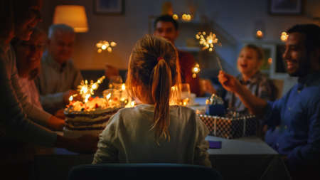 Back View Shot: Birthday Celebration of Cute Little Girl at the Dinner Table Blowing Out Candle on Her Cake. At the Table Happy Relatives and Friends Gathered To Celebrate.の写真素材