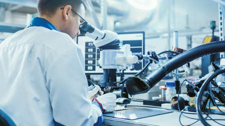 Electronics Factory Worker in White Work Coat is Soldering a Printed Circuit Board Through a Digital...