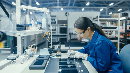 Young Female in Blue Work Coat is Assembling Printed Circuit Boards for Smartphones. Electronics...