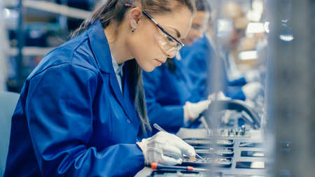 Female Electronics Factory Workers in Blue Work Coat and Protective Glasses Assembling Printed Circuit Boards for Smartphones with Tweezers. High Tech Factory with more Employees in the Background.の写真素材