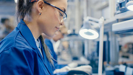 Woman Electronics Factory Worker in Blue Work Coat and Protective Glasses is Assembling Smartphones with Tweezers and Screwdriver. High Tech Factory Facility with more Employees in the Background.の写真素材