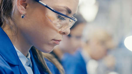 Close-up Shot of a Female Electronics Factory Worker in Blue Work Coat and Protective Glasses is...