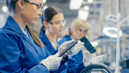 Woman Electronics Factory Worker in Blue Work Coat and Protective Glasses Takes a Smartphone Screen and Performs a Quality Check. High Tech Factory Facility with Multiple Employees.の写真素材