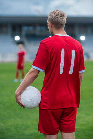Back Shot of a Professional Football Player Standing on a Stadium during the Championship Game and Holding a White Football Ball. His Teammate is in the Background.の写真素材