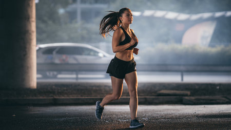 Shot of a Beautiful Fitness Girl in Black Athletic Top and Shorts Jogging in a Dark Foggy Street. She is Running in an Urban Environment Under a Bridge with Cars in the Background.の写真素材