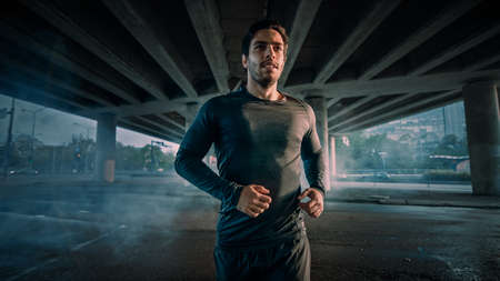 Portrait Shot of an Athletic Muscular Young Man in Sports Outfit Jogging in the Street. He is Running in an Urban Environment Under a Bridge.の写真素材