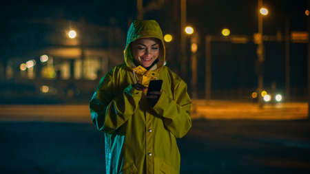 Beautiful Young Girl in a Yellow Raincoat is Using a Smartphone in the Rain. Shes Smiling After Receiving a Notification. Weather is Rainy and its Night Time.の写真素材