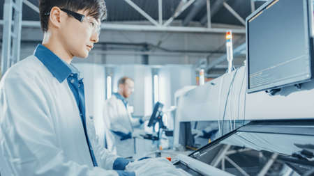 On High Tech Factory Asian Engineer Uses Computer for Programing Pick and Place Electronic Machinery for Printed Circuit Board Assembly Line. Production of PCB with SM Machinery.の写真素材