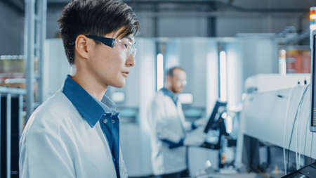 Portrait Shot of an Asian Engineer in High Tech Factory Uses Computer for Programing Pick and Place Electronic Machinery for Printed Circuit Board Assembly Line. Production of PCB with SM Machinery.の写真素材