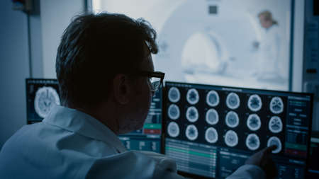 In Medical Laboratory Patient Undergoes MRI or CT Scan Process under Supervision of Radiologist, in Control Room Doctor Watches Procedure and Monitors with Brain Scans Results.の写真素材
