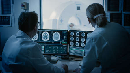 In Control Room Doctor and Radiologist Discuss Diagnosis while Watching Procedure and Monitors Showing Brain Scans Results, In the Background Patient Undergoes MRI or CT Scan Procedure.の写真素材