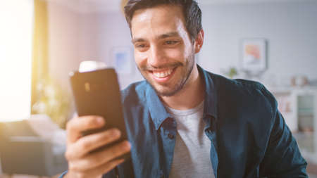 Handsome Smiling Man Uses Smartphone while Relaxing in His Living Room. Guy Browsing in Internet,...