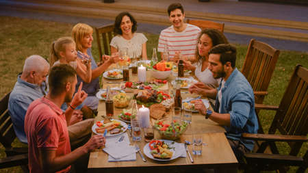 Group of Friends Gathered Together at the Table Relatives and Friends, Young and Elderly are Eating, Drinking, Passing Dishes, Joking and Having Fun. Garden Party Evening Celebration.の写真素材