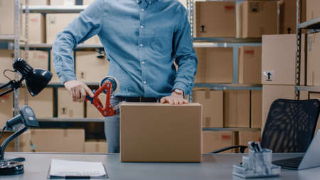 Professional Warehouse Worker Checks and Seales Cardboard Box Ready for Shipment. In the Background Person Working in the Rows of Shelves with Cardboard Boxes with Ready Orders.の写真素材