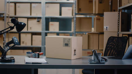 Cardboard Box Package Standing on the Table of the Warehouse where Rows of Shelves with Parcels Waiting to be Shipped and Delivered.の写真素材