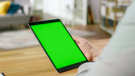 Man Using Green Mock-up Screen Digital Tablet Computer in Portrait Mode while Sitting at His Desk. In the Background Cozy Living Room.の写真素材