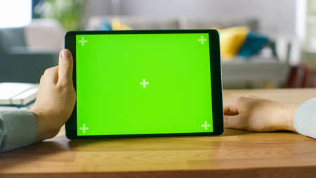 Man Holding and Watching Green Mock-up Screen Digital Tablet Computer While Sitting at the Desk. Man Watching Videos in Landscape Mode.の写真素材