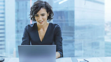 Portrait of the Successful Smiling Businesswoman Working on a Laptop in Her Office with Cityscape...