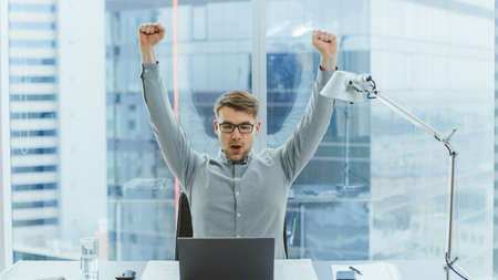 Handsome Young Businessman Sitting at His Desk Using Laptop in the Office Raises Hands and...