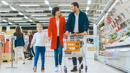 At the Supermarket: Happy Family of Three, Holding Hands, Walks Through Fresh Produce Section of the Store. Father, Mother and Daughter Having Fun Time Shopping.の写真素材