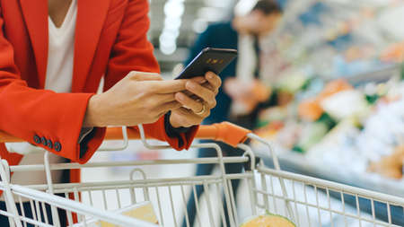 At the Supermarket: Woman Uses Smartphone, Leans on the Shopping Cart. In the Big Mall Woman Browsing In Internet on Her Mobile Phone. Focus on Hands Holding Mobile Phone.の写真素材