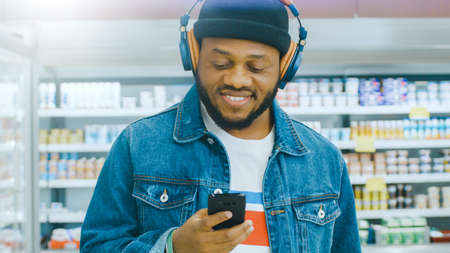 At the Supermarket: Portrait of a Stylish African American Guy with Headphones, Using Smartphone,...