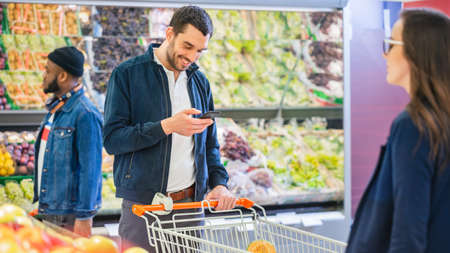At the Supermarket: Handsome Man Uses Smartphone while Standing in the Fresh Produce Section of the...