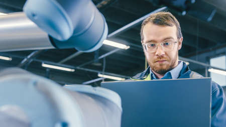 Low Angle Shot At the Factory: Automation Engineer Uses Laptop for Programming and Testing Robotic...
