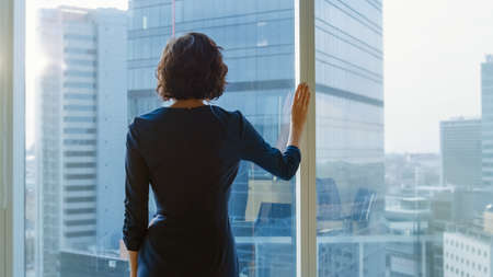 Shot of the Successful Businesswoman in a Striking Black Dress in Her Office Looking out of the Window Thoughtfully. Modern Business Office with Personal Computer and Big City View.の写真素材