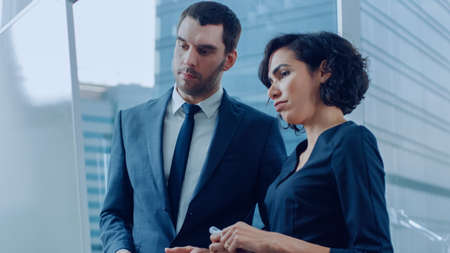 In the Office Handsome Businessman and Beautiful Businesswoman Think About Future Plans Written on the Whiteboard. Planning Marking Expansion, Consulting Data and Making Graphs. Modern Stylish People.の写真素材