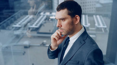 Confident Businessman in a Suit Standing in His Office, Looking out of the Window and Making Important Phone Call so He can Close the Deal. In the Background View with Big City.の写真素材