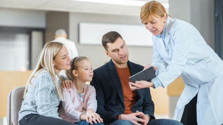 In the Hospital Lobby Female Doctor Shows Information on the Tablet Computer to the Young Family...