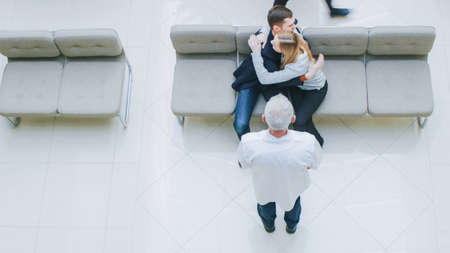 High Angle Shot in the Hospital, Young Couple Sitting in the Lobby and Hugging. They Receive Test Results with Good News from their Doctor. Happy Moment in the Life of Young People.の写真素材