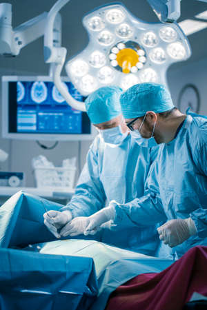 Portrait of the Professional Surgeons Performing Invasive Surgery on a Patient in the Hospital Operating Room. In the Background Modern Hospital Operating Room. Vertical Shot.の写真素材