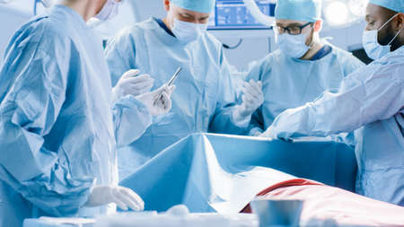Diverse Team of Professional surgeon, Assistants and Nurses Performing Invasive Surgery on a Patient in the Hospital Operating Room. Nurse Hands Out Instruments. Real Modern Hospital with Equipment.の写真素材
