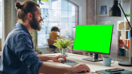 Handsome Long-Haired Bearded Manager Working at a Desk in Creative Office, Using Desktop Computer with Green Screen Mock Up Display. Colleagues Working in the Backgroundの写真素材