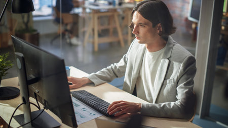 Stylish Long-Haired Manager Wearing a Gray Jacket, Working at a Desk in Creative Office. Young Handsome Man Using Desktop Computer in Marketing Agency. Colleagues Workingの写真素材