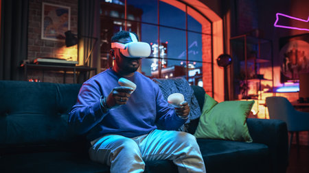Cinematic Portrait of a Young Stylish Black Man Using Virtual Reality Headset with Joysticks. African American Man Sitting on a Sofa at Home in the Evening, Playingの写真素材