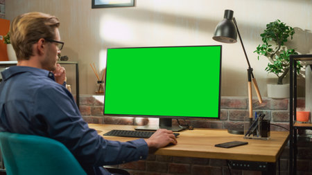 Young Handsome Man Working from Home on Desktop Computer with Green Screen Mock Up Display. Creative Male Checking Social Media, Browsing Internet. Living Room inの写真素材