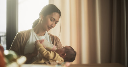 Portrait of Beautiful Asian Mother Feeding Milk to her Infant Using a Baby Bottle at Bright Home. Young Woman New to Motherhood Bonding with her Child and Enjoying anの写真素材