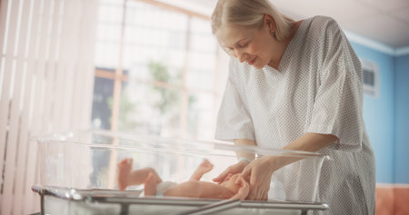Mother Bonding with her Newborn Child Near Hospital Bassinet Bed in a Modern Nursery Clinic. Caring...