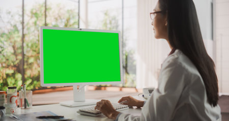 Young Female Doctor Wearing White Coat, Working on Personal Computer with Green Screen Mock Up Display in Her Office. Beautiful Asian Medical Health Care Professional Workingの写真素材
