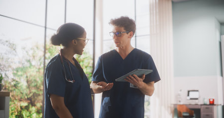 Two Young Doctors Standing in a Medical Facility in the Hospital, Talking and Sharing Information on Tablet Computer. Modern Hospital with Diverse Multiethnic Professionalの写真素材