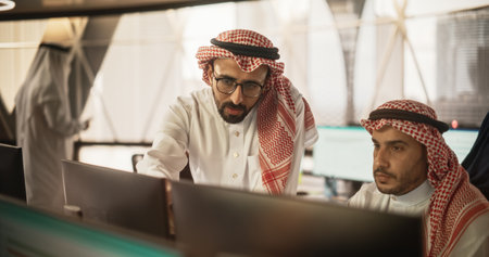 Arab Graphic Designer Working on Computer, Discussing Project with Team Leader. Middle Eastern Colleagues Working in Modern Office, Wearing Traditional Whiteの写真素材
