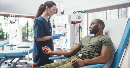 Caucasian Female Nurse Taking Blood Donation From Black Army Soldier In Military Hospital. African American Man Squeezing Heart-Shaped Ball To Pump Blood Into Bag. Troop Donatingの写真素材