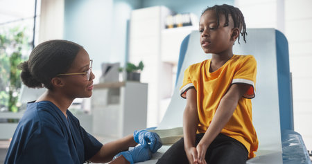 African Boy Sitting In The Chair In Hospital, Talking To Female Black Nurse. Professional Woman Listening To Young Man's Symptoms, Gives Advice On Proper Treatment.の写真素材